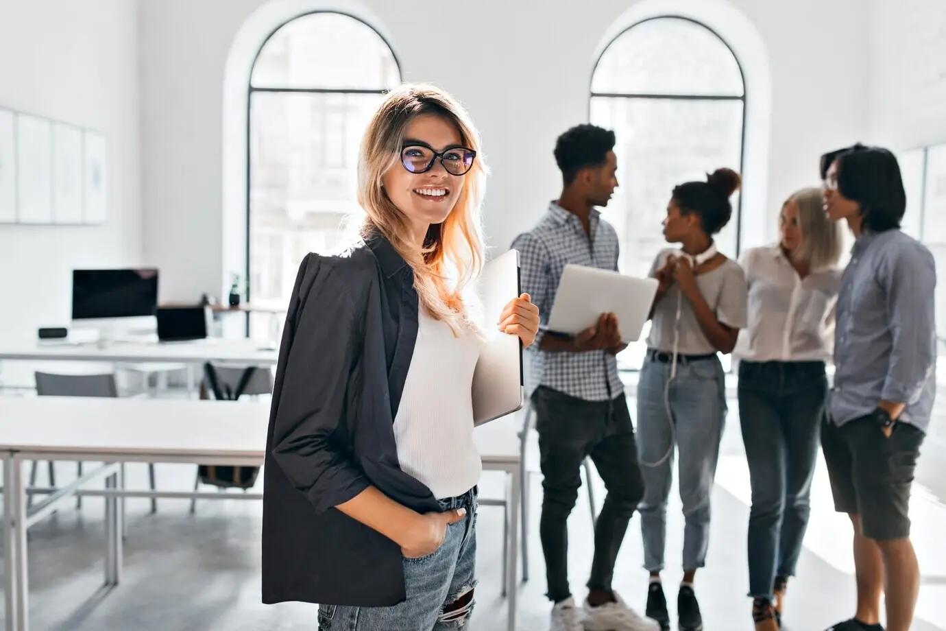 Indoor portrait of an elegant businesswoman in a black jacket with her team. An African office manager in white sneakers carries a laptop and talks with a mixed-race woman in jeans.