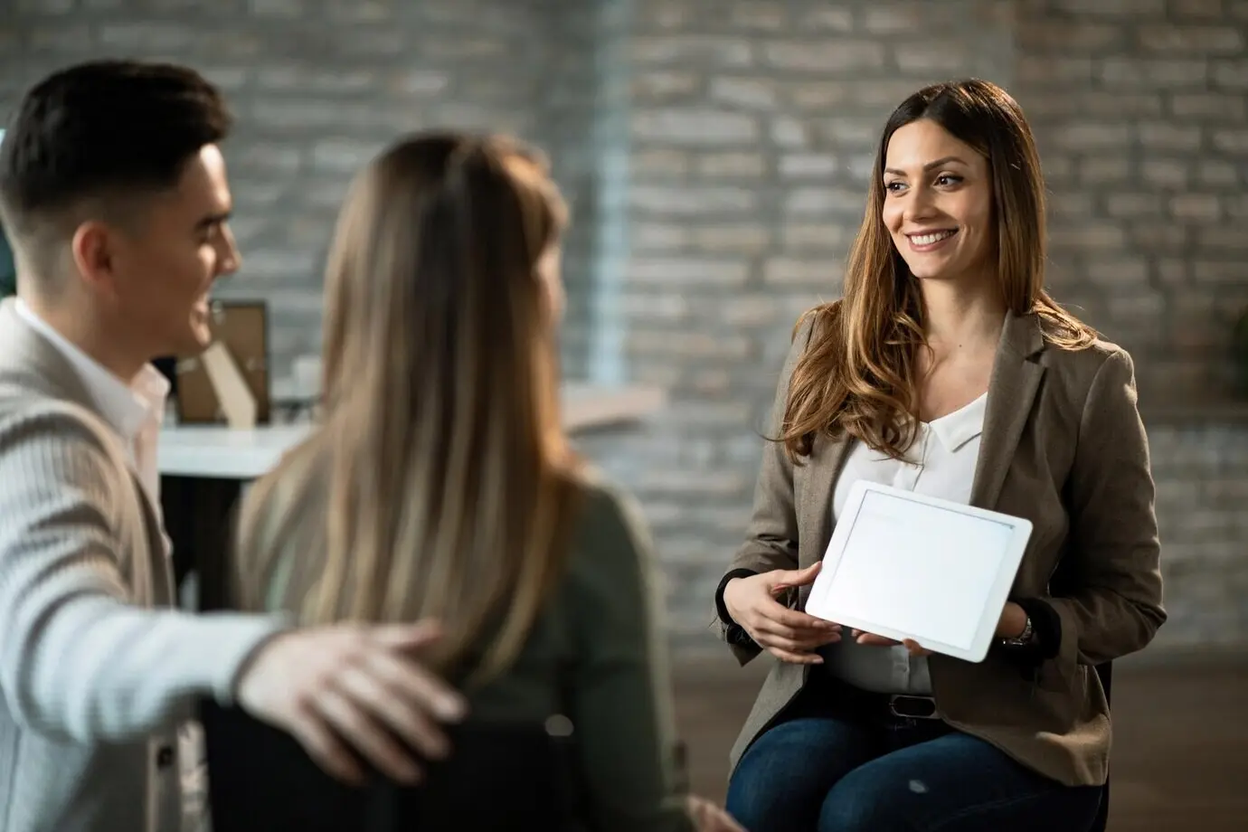 A smiling financial advisor meets with a young couple in an office, showing them investment options on a touchpad.